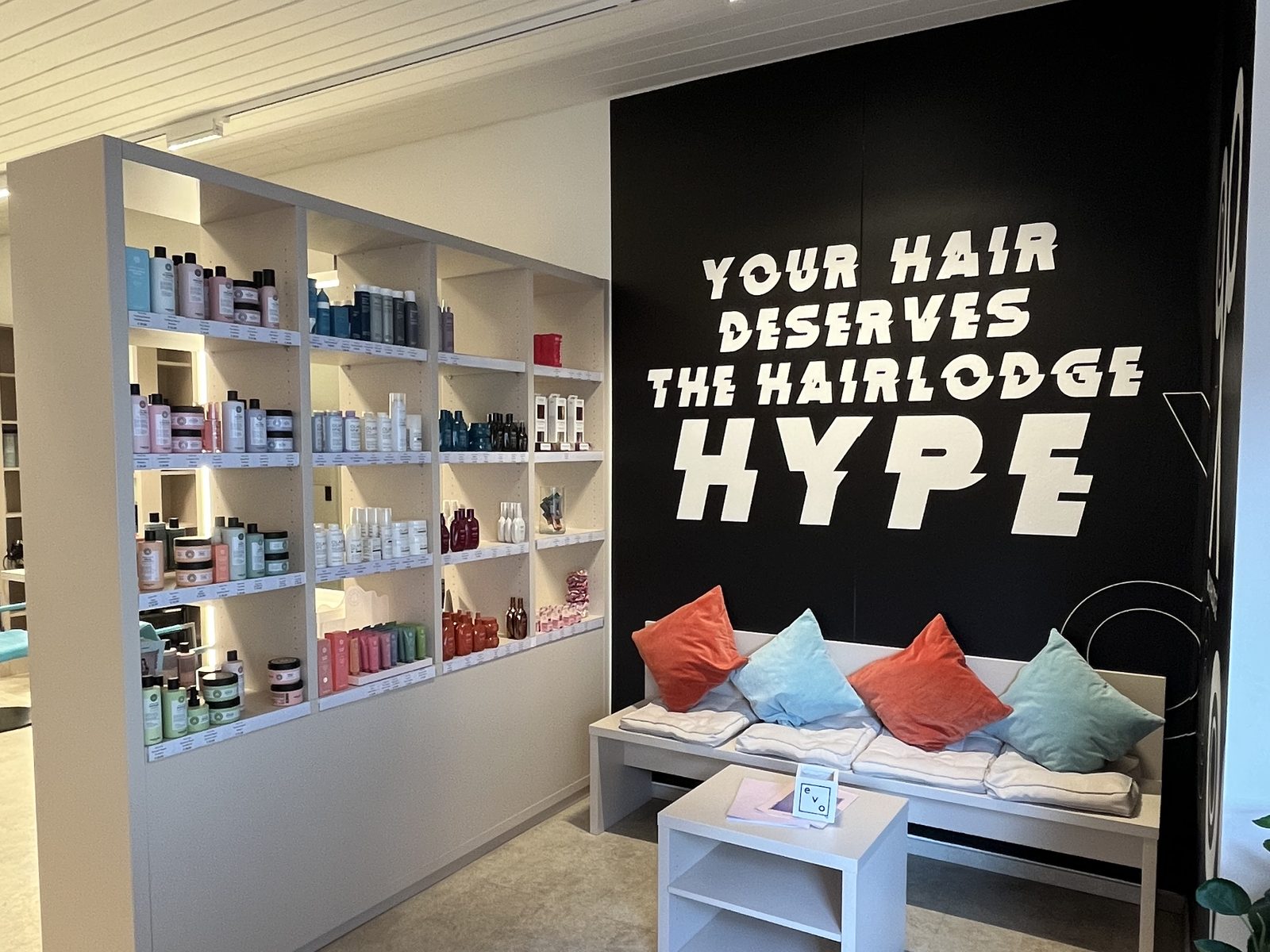 Waiting area at HAIRLODGE Ellmau: a pale bench with colourful cushions in front of the black wall reading "Your hair deserves the Hairlodge Hype", next to a shelf full of hair-care products.
