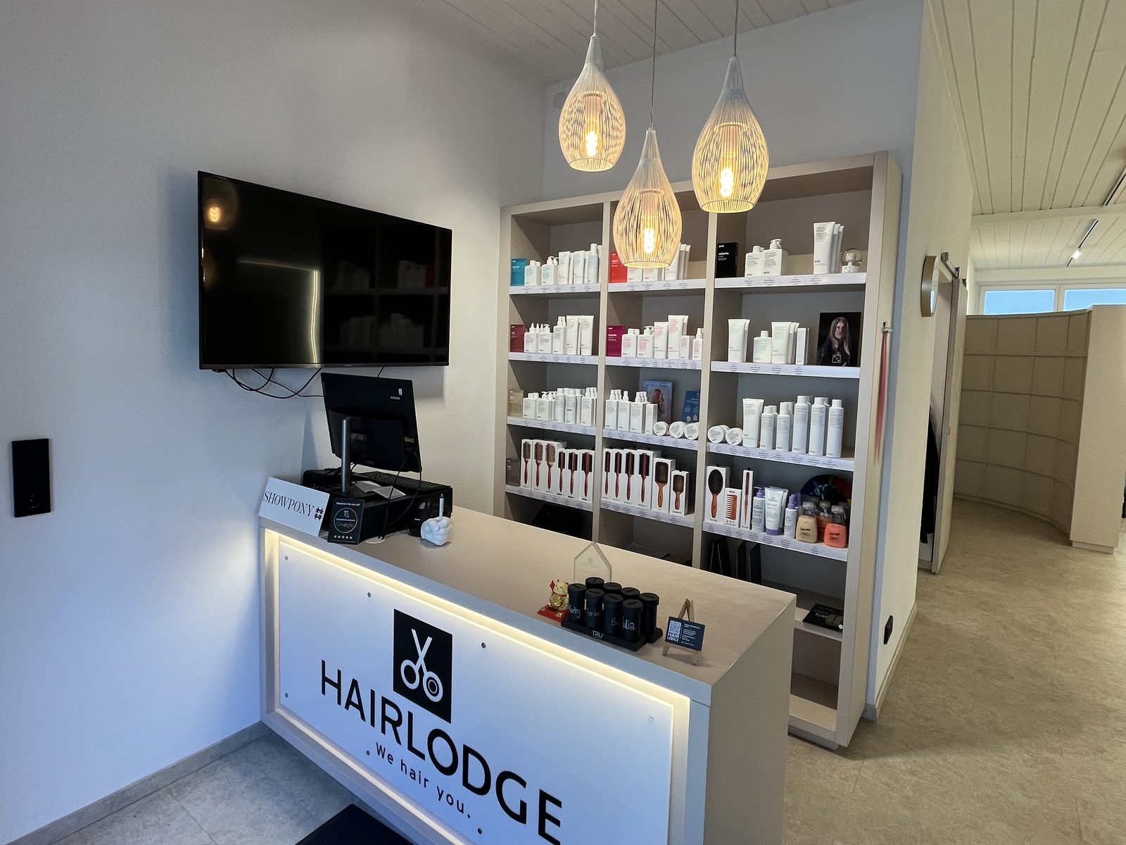 Illuminated HAIRLODGE reception desk at the Ellmau salon, with three pendant lamps above and a shelf of care and styling products behind.