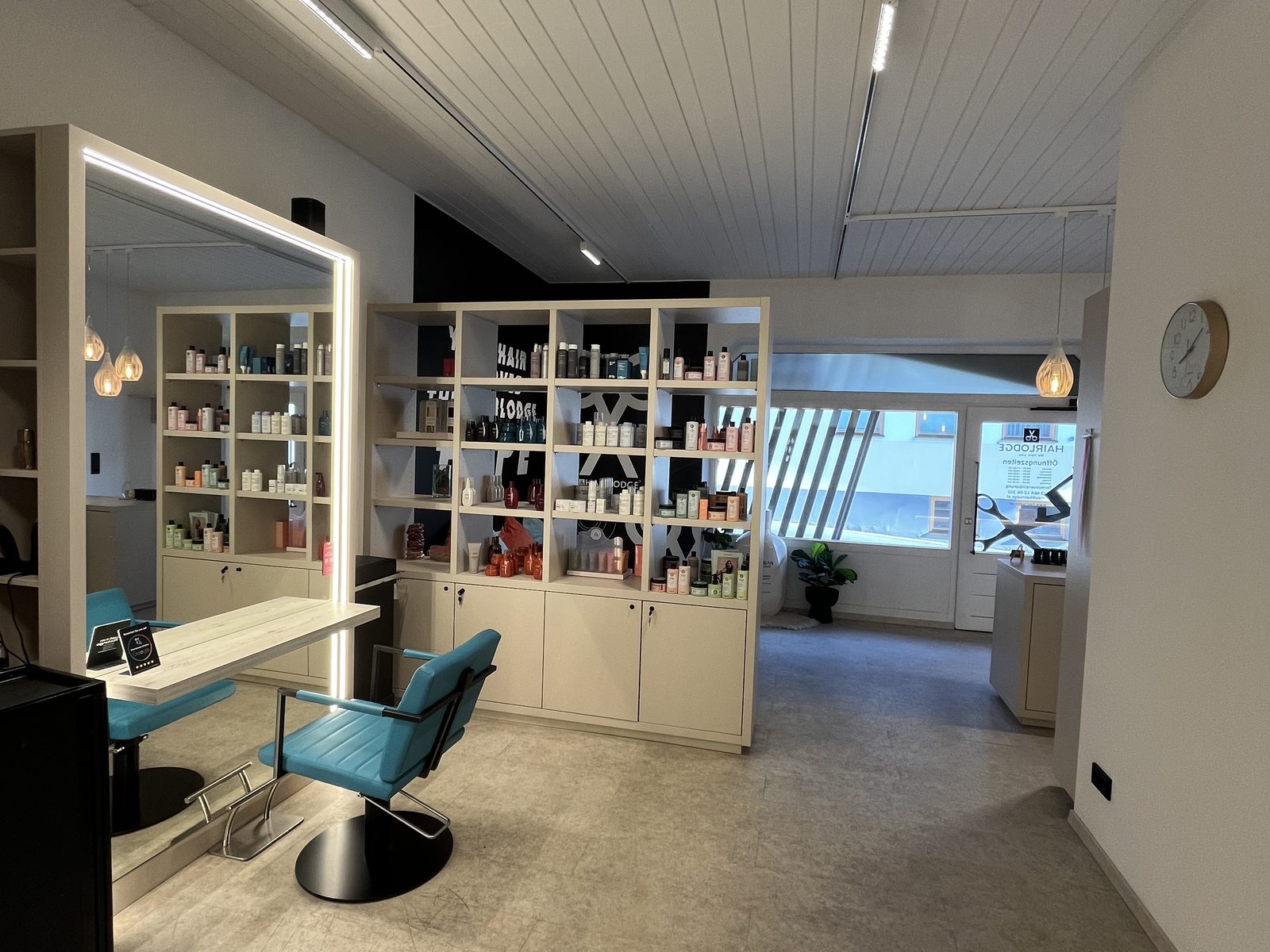 Wide view through HAIRLODGE Ellmau towards the entrance, with product shelving, a styling station and the wall clock.