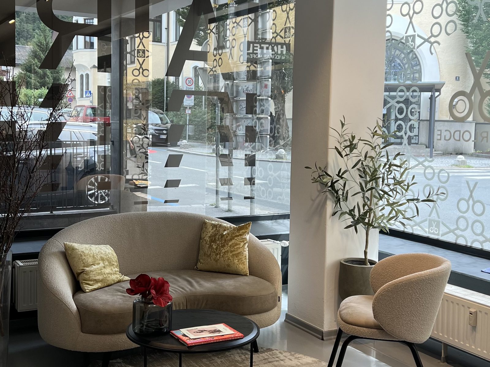 Waiting area at HAIRLODGE Kufstein with a curved sofa, armchair and olive tree in front of the floor-to-ceiling windows.