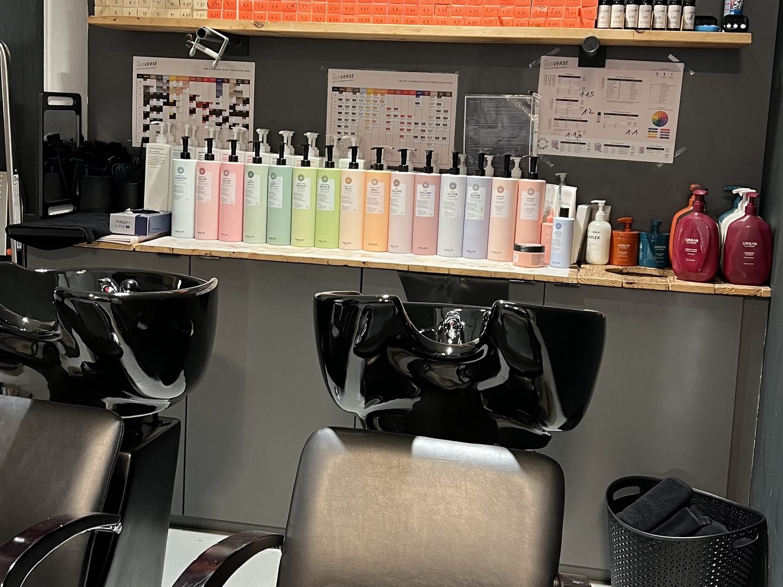 Wash area at HAIRLODGE Kufstein with black basins in front of the wooden shelf wall of care products.