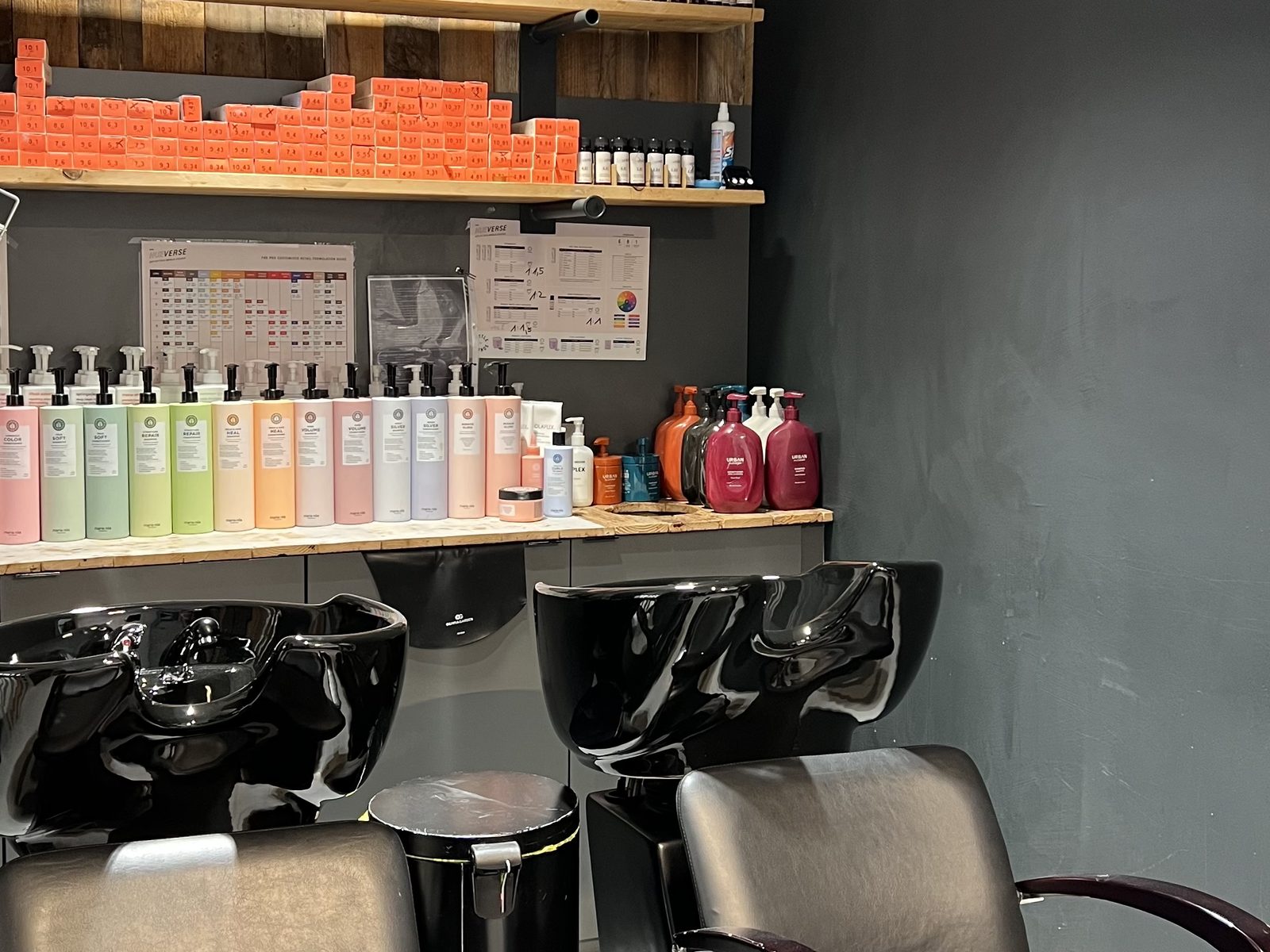 Wash stations at HAIRLODGE Kufstein against the anthracite wall, with colour tubes and care products on the shelves above.