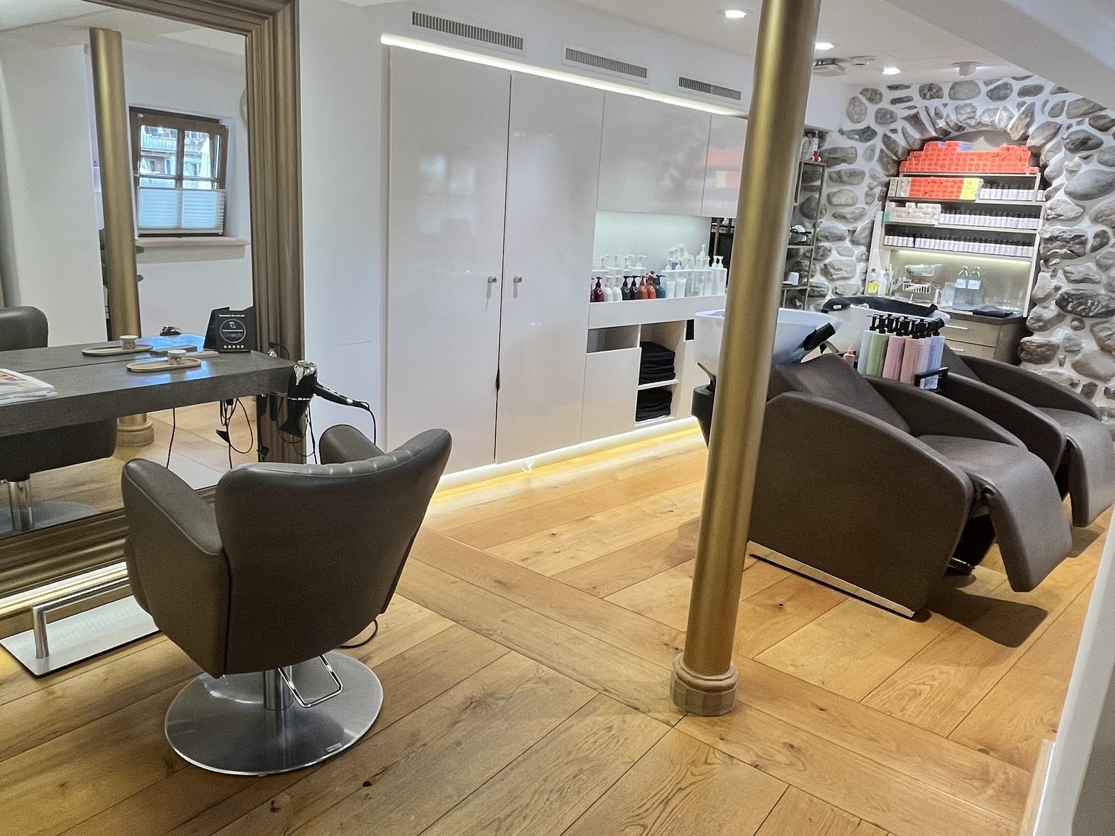 View into HAIRLODGE Stanglwirt: styling station with a large mirror, brass column and the washing area in front of the natural stone wall behind it.